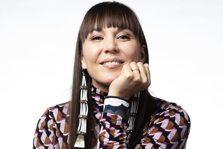 Woman with long brown hair and bangs, wearing a patterned shirt and long earrings, smiling with her chin resting on her hand.