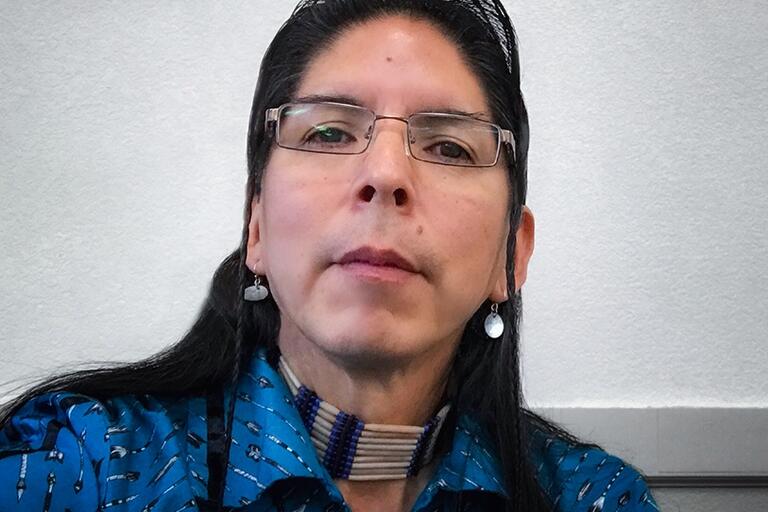 A man with long dark hair wears glasses, beaded jewelry, and a blue shirt