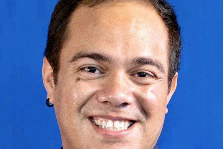 Close-up of a smiling person with short dark hair against a blue background.