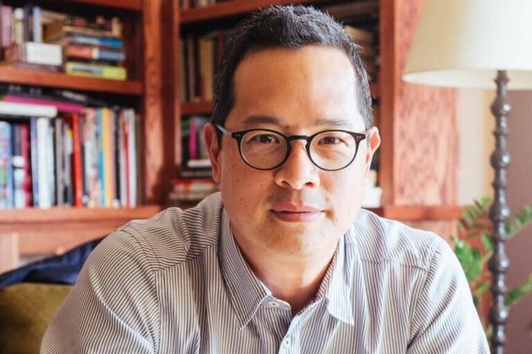 Man with black-rimmed glasses in a striped shirt sitting in front of a bookshelf filled with books.
