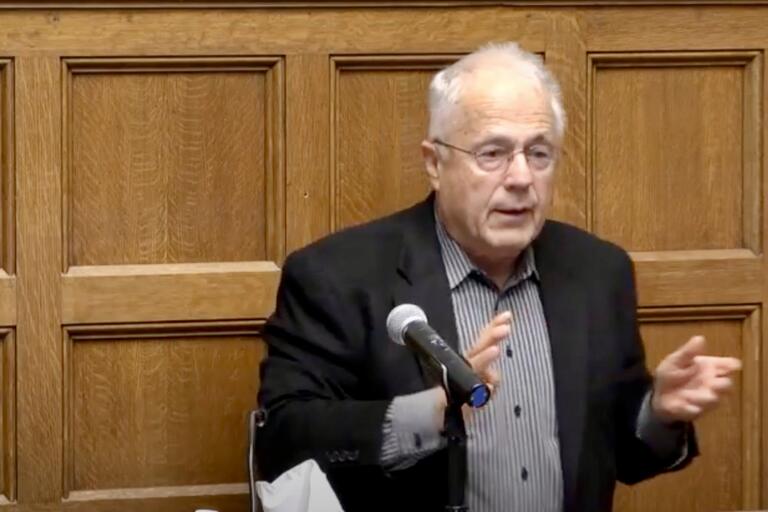 An older man speaking at a podium with a wooden panel wall background.