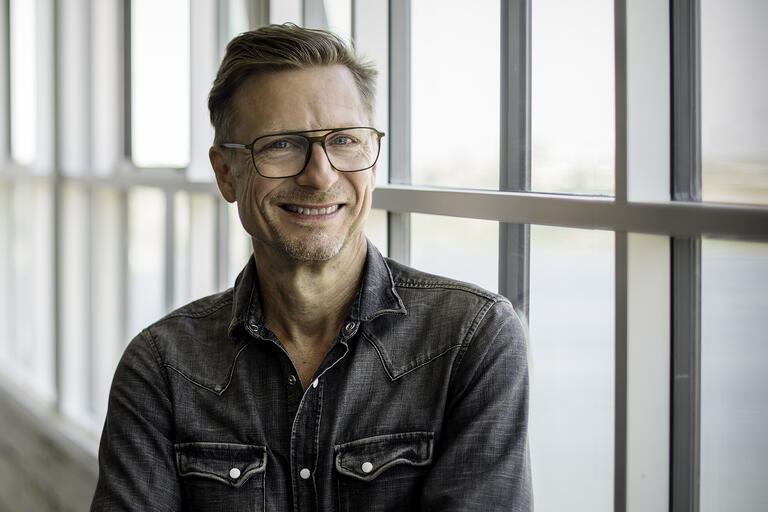 Man wearing glasses and a dark denim shirt, standing in front of a large window.