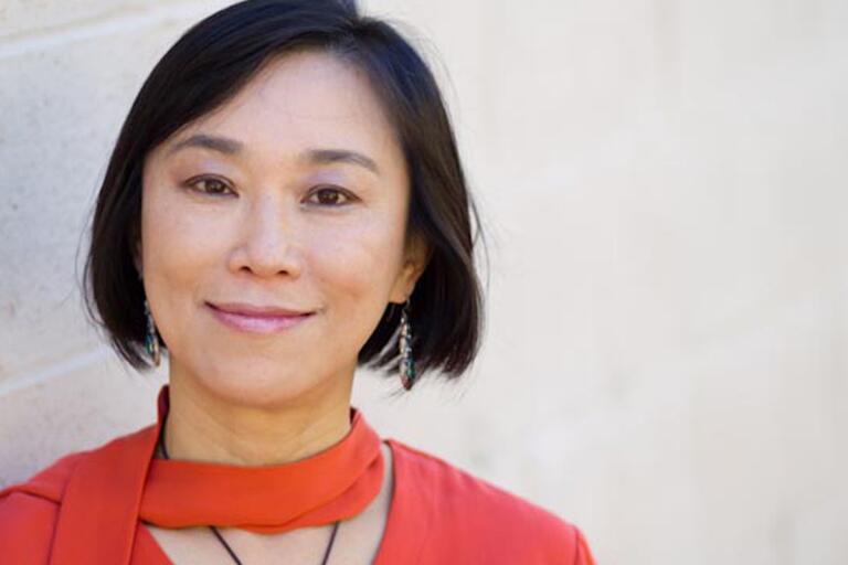A woman with short black hair, wearing a red top and scarf, standing against a beige brick wall, smiling.
