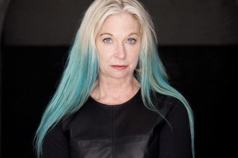 Woman with blonde and teal hair in a black top, standing with arms crossed.