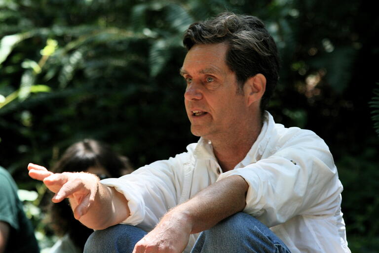 Man sitting outdoors, gesturing with his right hand, wearing a white shirt and blue jeans.