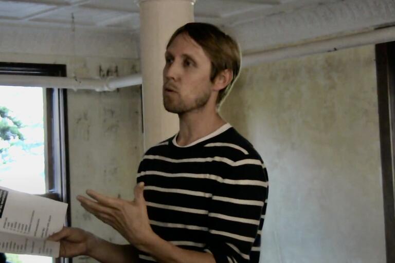 Man in a striped shirt holding a document and speaking indoors.