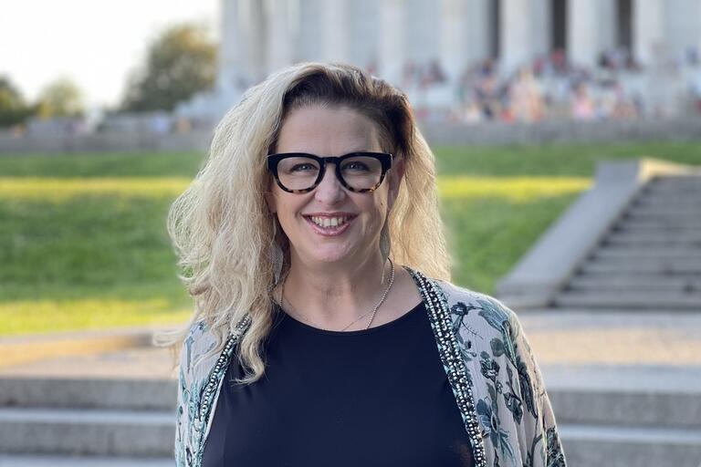 Person with blonde hair and black-rimmed glasses smiling in front of stone steps and a large building.