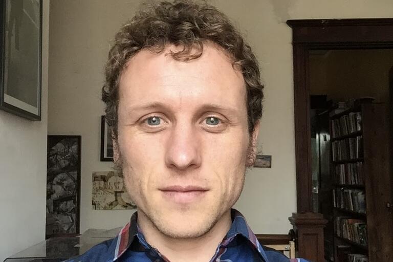 Close-up of a person with curly hair, indoors with artworks and bookshelves in the background.
