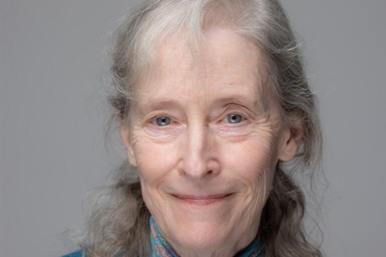 eadshot of an elderly woman with long gray hair and blue eyes, wearing a turquoise top with a patterned scarf.