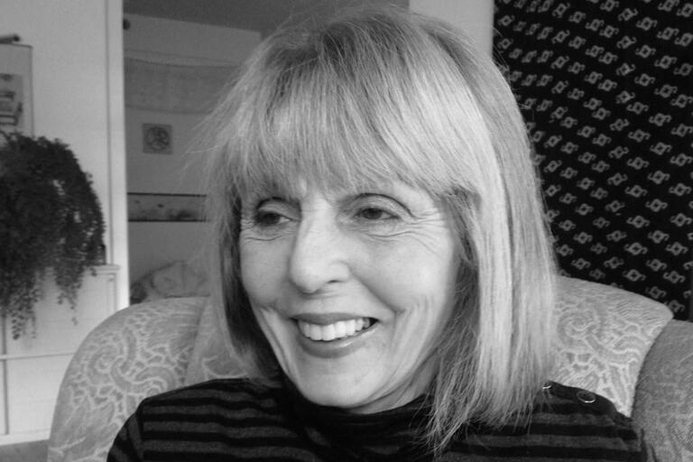 A smiling woman with chin-length hair and bangs, wearing a striped top, seated in a patterned chair.