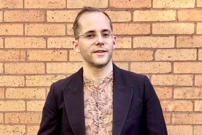 Person wearing a dark jacket and patterned shirt, standing in front of a red brick wall.