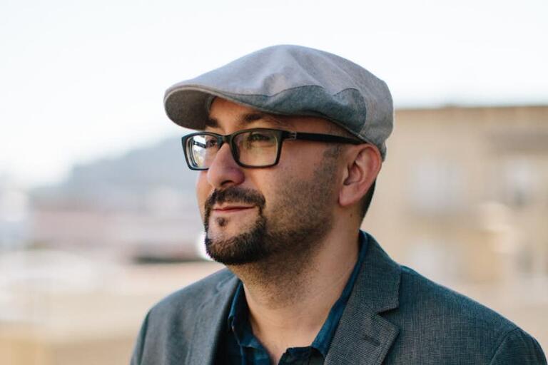 Person wearing a light grey flat cap, black-framed glasses, and a charcoal grey blazer over a dark blue collared shirt, smiling slightly and looking to the right.