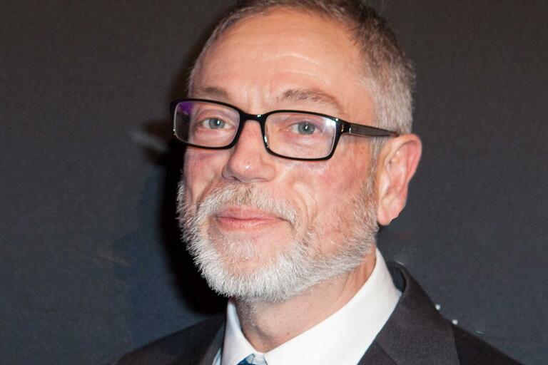 Close-up of an older man with gray hair, beard, and glasses, wearing a suit and tie.