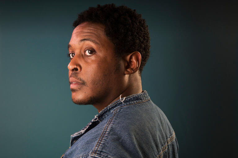 Close-up side profile of a person with short curly hair and a denim jacket, facing left with a direct gaze.