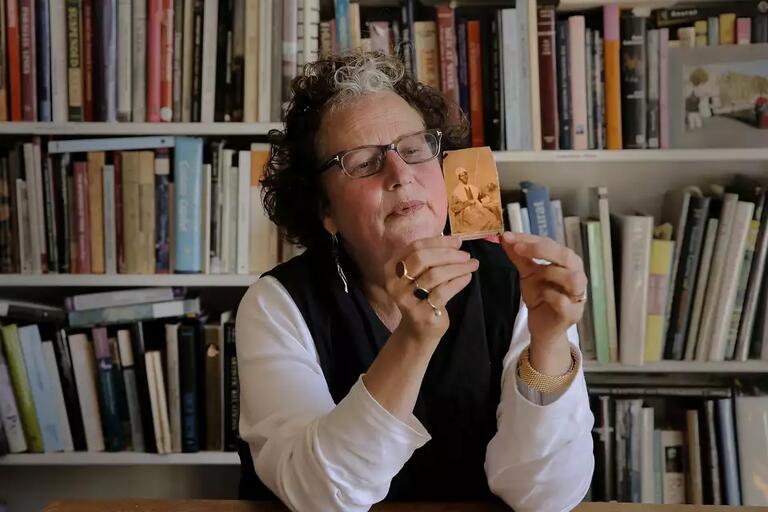 Elderly person with glasses holds a small sepia photograph, seated in front of a bookshelf filled with various books.