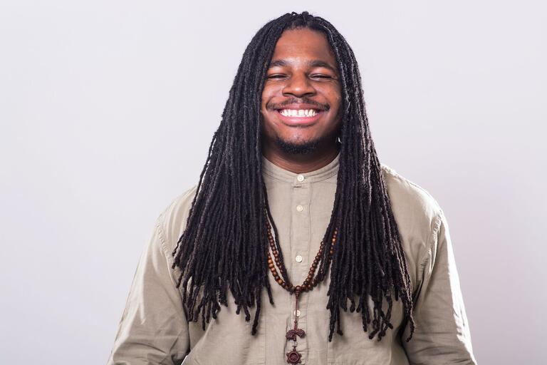Person with long dreadlocks, wearing a beige shirt and brown bead necklaces, smiling with eyes closed.
