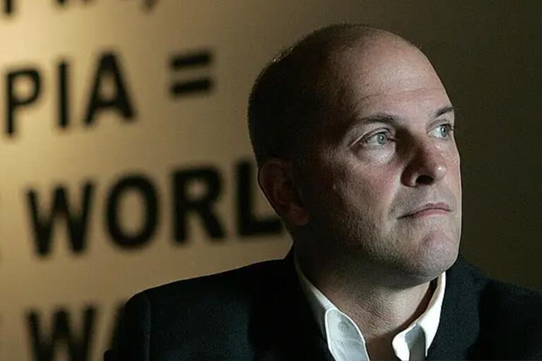 A person in a dark suit and white shirt looks off-camera against a blurred text background.