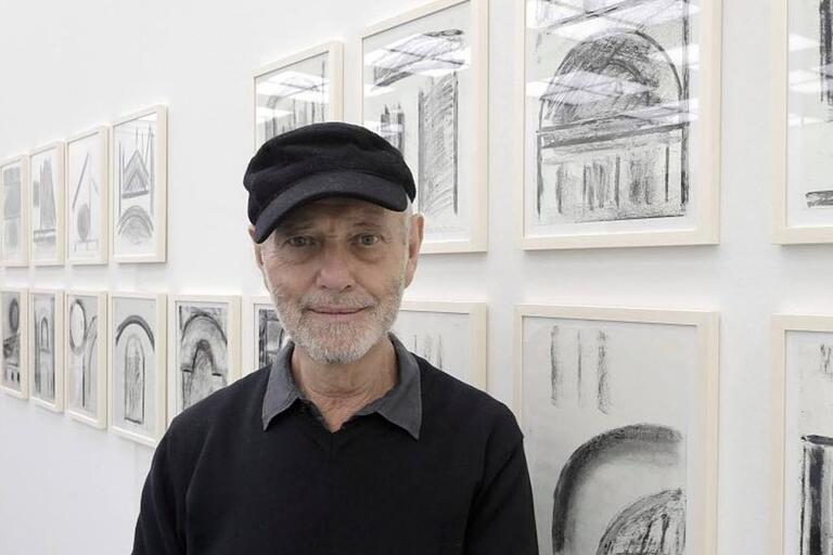 "Elderly man with a black cap standing in front of a wall of framed architectural sketches."