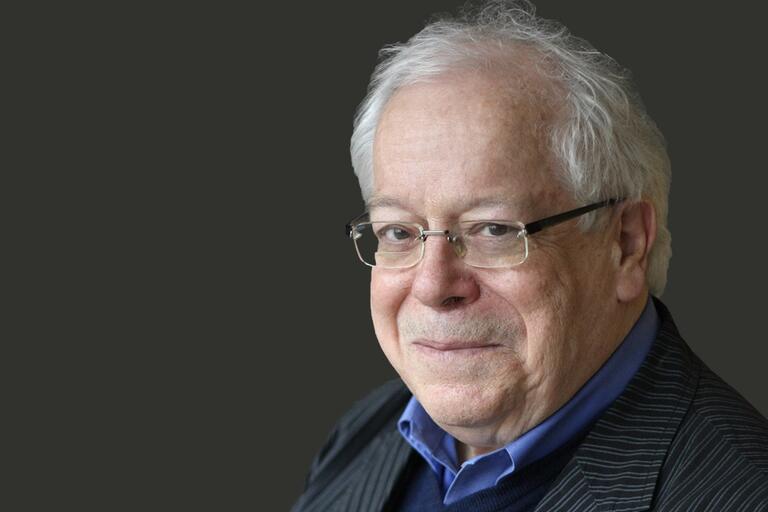 Portrait of an older man with white hair and glasses dressed in professional attire against a dark grey background.