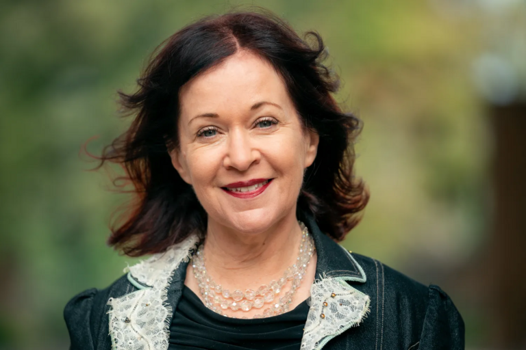 Portrait of a middle-aged woman smiling, wearing a black jacket with lace trim and a multi-layered pearl-like necklace, with a blurred green background.