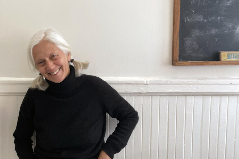 Person with white hair and black turtleneck smiling in front of a white wall and chalkboard.