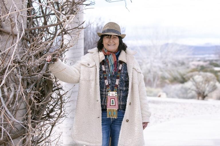 Person in a beige winter coat and hat standing outdoors near a bare tree.