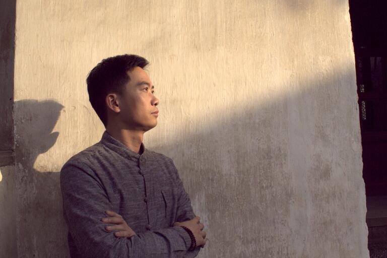 A man with crossed arms leans against a textured beige wall, casting a shadow.