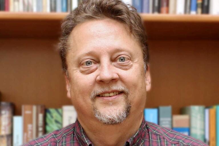 iddle-aged man with blue eyes and a beard smiling in front of a bookshelf filled with books.