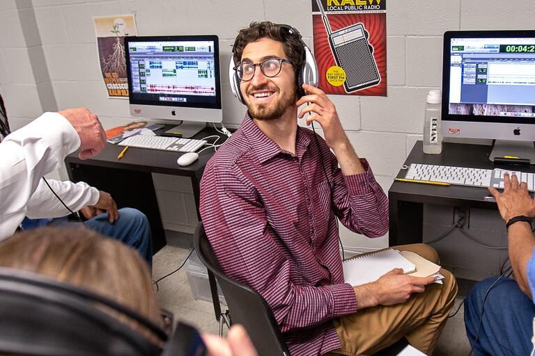 Three people working in a radio studio, central figure wearing headphones and smiling at a computer with audio editing software, KALW radio poster on the wall.