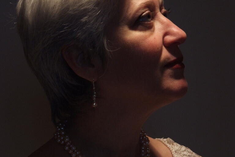 Profile of an older woman with gray hair, pearl earrings, and a lace top, illuminated against a dark background.