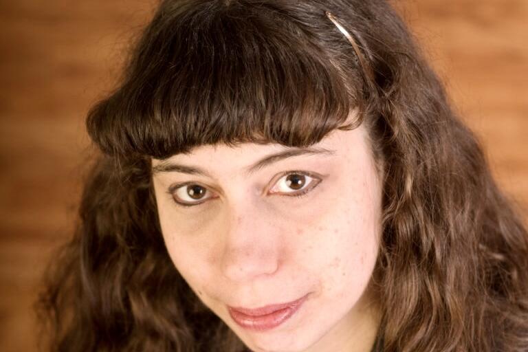 Close-up portrait of a person with long, wavy dark brown hair, bangs, and brown eyes. They have a neutral expression and are wearing a metallic hair clip. The background is a brown gradient.