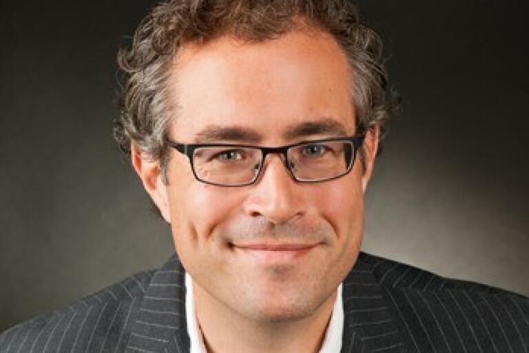 Headshot of a middle-aged man with short, wavy, brown hair and black glasses, wearing a white shirt and dark pinstriped blazer against a dark gradient background.