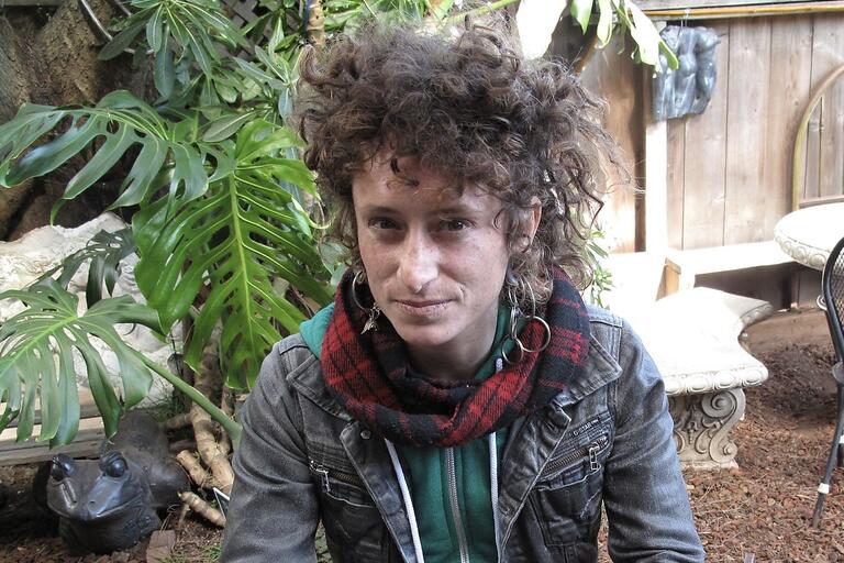 Person with curly hair in a garden wearing a denim jacket, green hoodie, and red and black plaid scarf.
