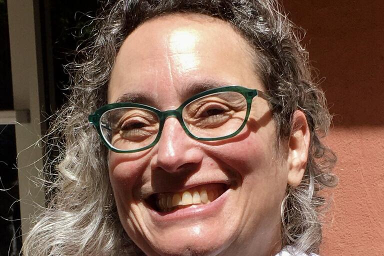 Close-up of a smiling person with green-framed glasses and curly, salt-and-pepper hair.