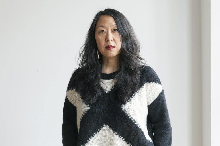 Woman with long dark hair wearing a black and cream X-patterned sweater standing against a plain light background.