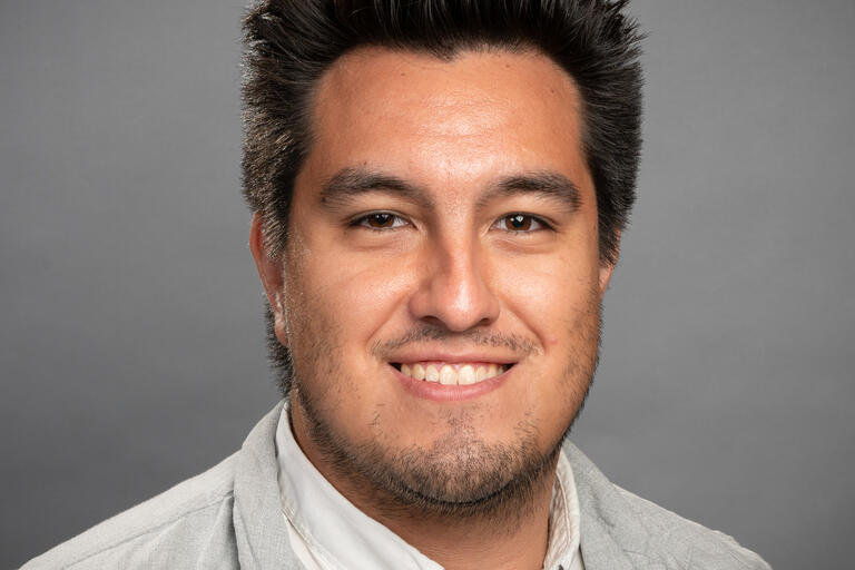 Close-up portrait of a person with dark, styled hair, light stubble, and a slight smile, wearing a light-colored collared shirt, against a plain grey background.