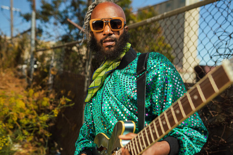 Person wearing a sequined jacket and sunglasses, holding an electric guitar outdoors.