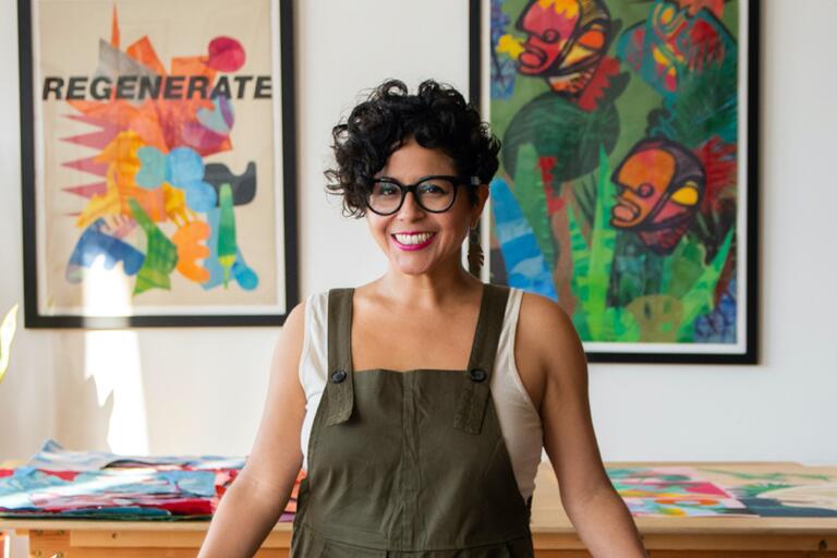 Person with short curly hair and glasses smiling, standing in front of colorful abstract artwork.