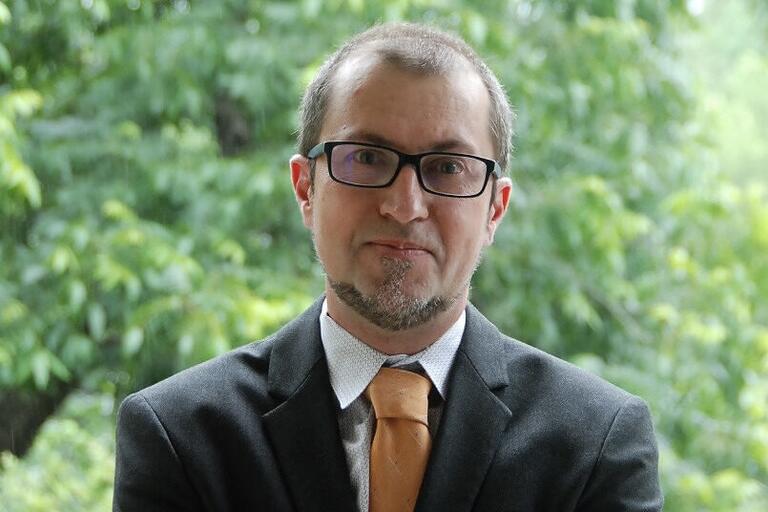 Person in a suit with an orange tie standing in front of a green foliage background.