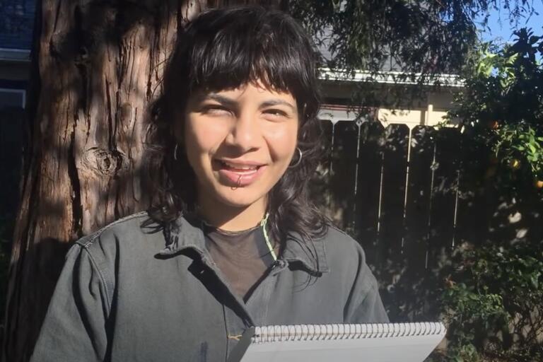 Person holding a notebook, standing in front of a tree in a garden, smiling at the camera.
