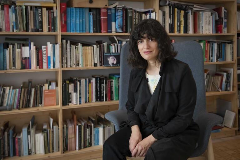 A woman with curly hair sits on a grey armchair in front of bookshelves filled with books.