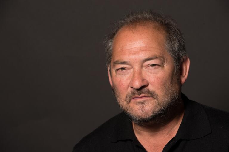 Headshot of a man with short, graying hair, a beard, and mustache, wearing a black collared shirt against a dark background.