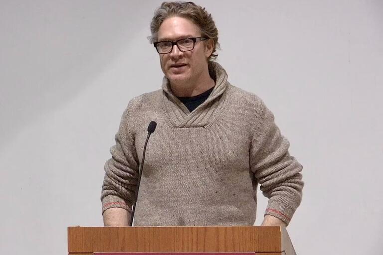 Man in a light brown sweater speaks at a wooden podium with a microphone.