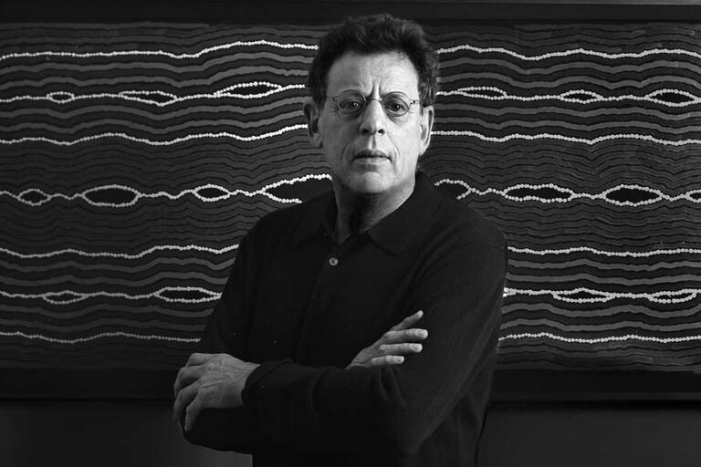 Black-and-white image of a man with crossed arms in front of abstract wavy artwork.