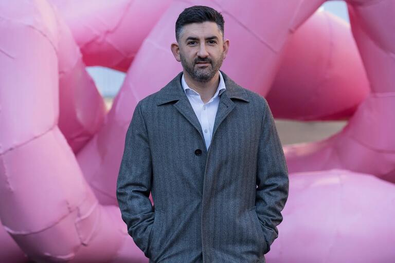 Man standing in front of a large pink inflatable art installation, wearing a gray coat and white shirt.