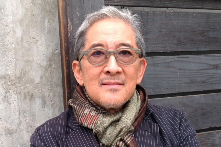 Older man with gray hair, glasses, and a multi-colored scarf, standing against a concrete and wooden wall background.