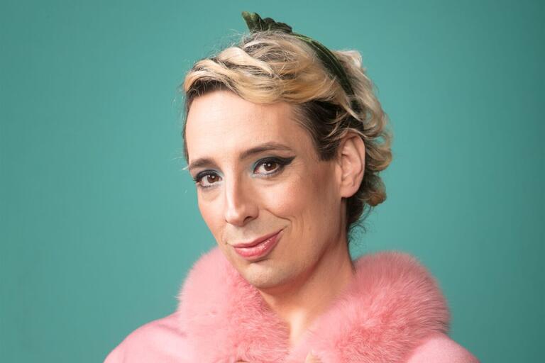 Headshot of a person smiling, wearing a pink garment with a fluffy collar and a dark green headband, set against a teal background.