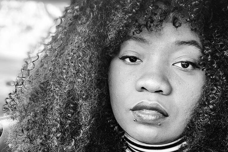 Close-up black-and-white portrait of a person with curly hair, eyeliner, two lip piercings, and a striped high-neck garment.