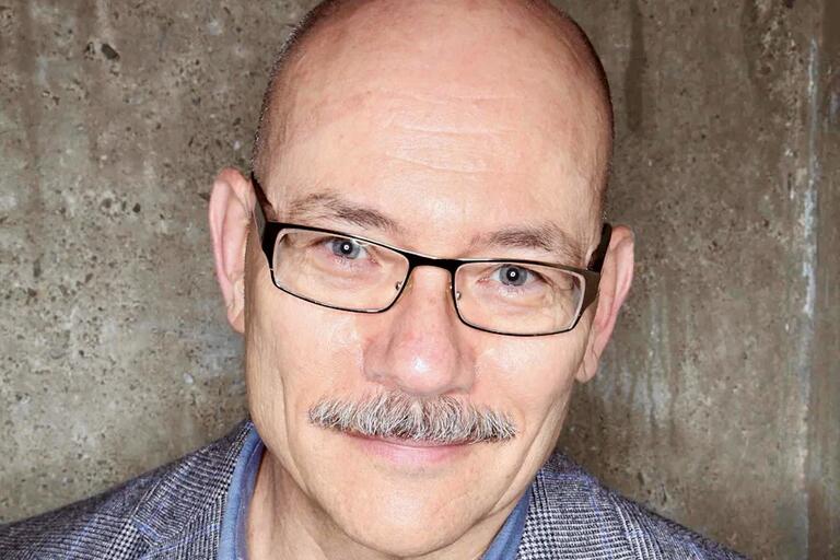 Close-up portrait of a bald man wearing glasses and a gray mustache against a concrete wall background.