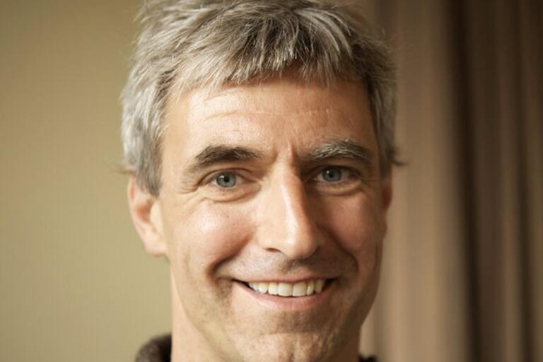 Close-up portrait of a smiling man with short gray hair and light blue eyes against a backdrop of neutral-colored drapes.